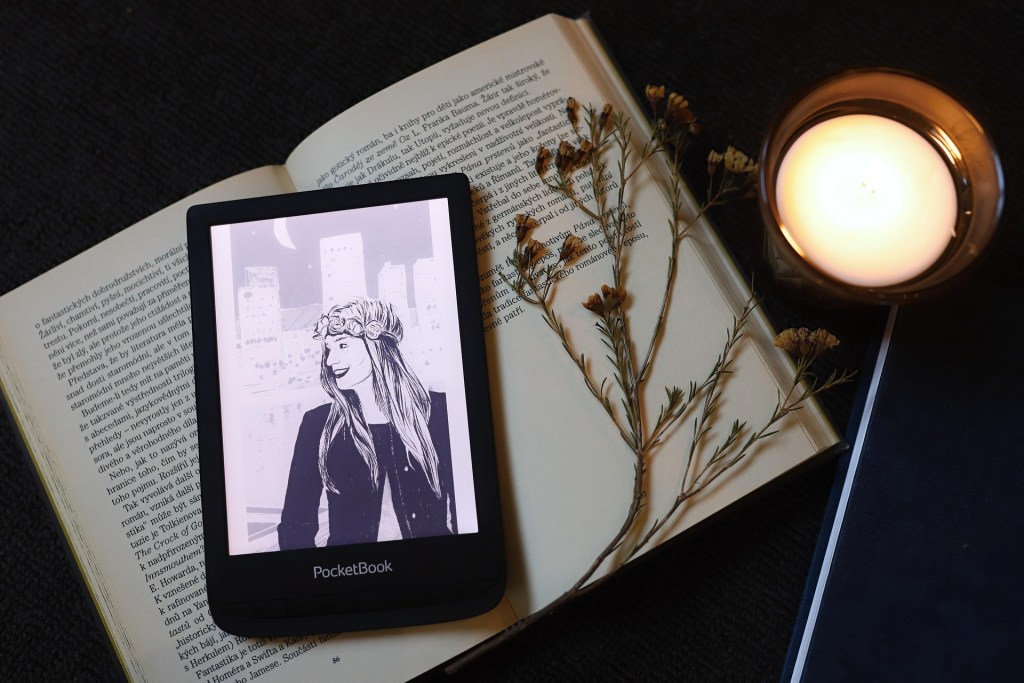 一個電子書閱讀器放在一本書上，螢幕顯示一位微笑的女子插畫，旁邊有一支乾花和一個點燃的蠟燭。
