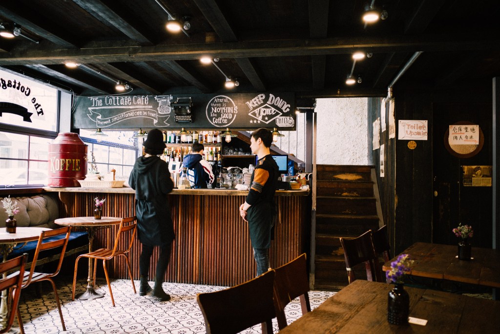 咖啡館內部，一名服務員在吧檯後準備飲品，另一名顧客正站在吧檯前聊天，周圍擺放著木製桌椅和花瓶，環境舒適且有特色