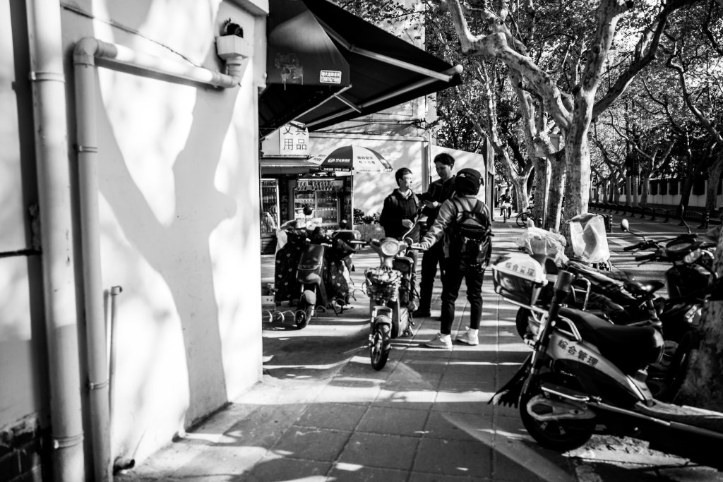兩名男子在街道上交談，旁邊有一輛電動車和數輛停放的摩托車，周圍環境有樹木和商店。