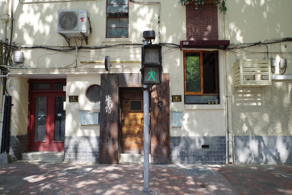 一棟矮樓的外觀，包含紅色和木質門，旁邊有行人信號燈和窗戶，周圍有陽光投射的樹影。